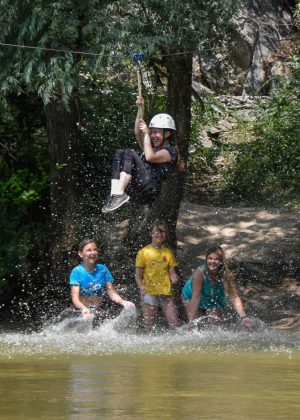 A girl doing zip-line above the river and three girls looking her smiling